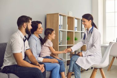Beyaz önlüklü bir kadın pediyatrist, klinikteki bir aileye danışıyor..
