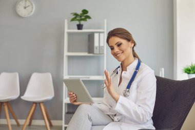 Doktor el sallıyor ve klinikte tablet kullanan bir hastayla görüntülü konuşma yapıyor..