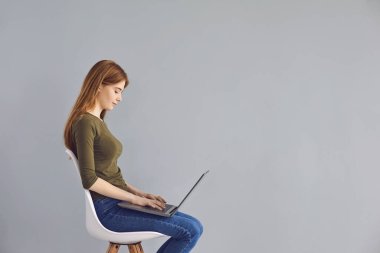 Dizüstü bilgisayarlı tatlı kızıl saçlı kız gri arka planda bir sandalyede oturuyor..
