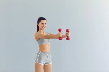 Gülümseyen genç kadın, gri arka planda dambıllarla antrenman yapıyor. Spor eğitmeni internette spor dersi veriyor.