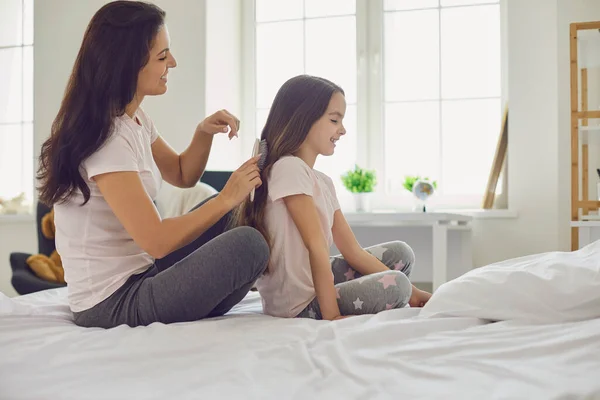 Ibu membuat gaya rambut untuk anak perempuan duduk di tempat tidur di kamar tidur. — Stok Foto