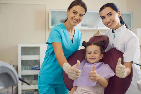 Dentista sonriente, asistente y niña pequeña paciente mostrando pulgares hacia arriba signo en la clínica dental —  Fotos de Stock