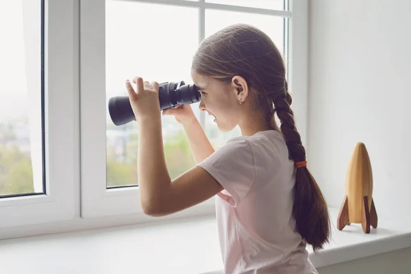 Konsep isolasi rumah selama epidemi pandemi. Seorang gadis dengan teropong tampak keluar jendela saat berdiri di ruangan. — Stok Foto