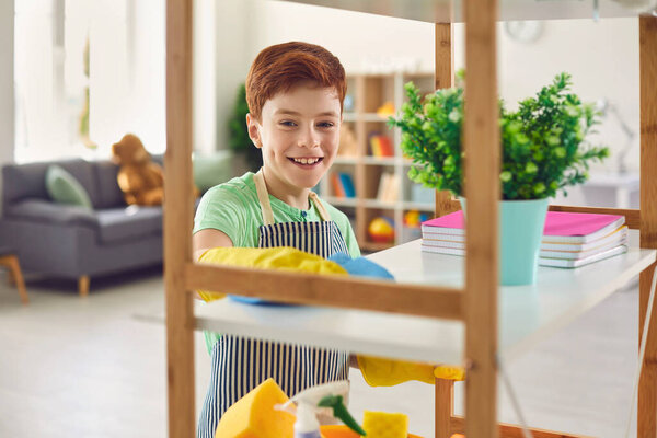 Adorable little boy in rubber gloves cleaning shelf with rag at home. Cute kid helping to do domestic chores