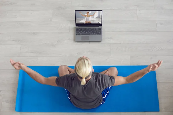 Meditación de yoga en línea. El rubio practica yoga en casa. Un tipo con el pelo blanco medita sentado en un piso gris en una habitación. — Foto de Stock