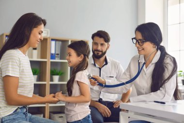 Aile doktoru, klinikteki masada oturan çocuğun steteskopuyla dinliyor..