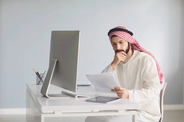 Orang Arab serius berpikir duduk di meja dengan komputer menganalisis melihat kertas. — Stok Foto