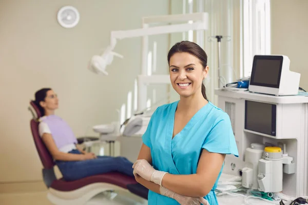 Smiling dentist standing and looking at camera with sitting patient at background