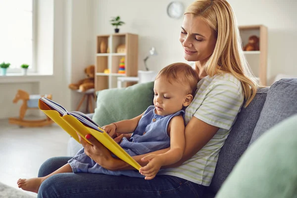 Mãe e criança estão lendo um livro enquanto estão sentados no sofá em casa. Dia das Mães. — Fotografia de Stock