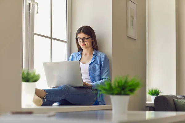 Young smiling woman sitting on windows and working online on laptop — Stock Photo, Image