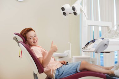 Diş sağlığı kliniği. Gülümseyen çocuk dişçi muayenesinde başparmak işareti gösteriyor ve kameraya bakıyor.