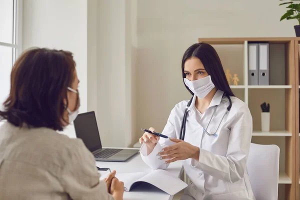 Doctora y paciente con máscaras protectoras hablando en la clínica. Cliente que recibe consulta de médico en el hospital —  Fotos de Stock