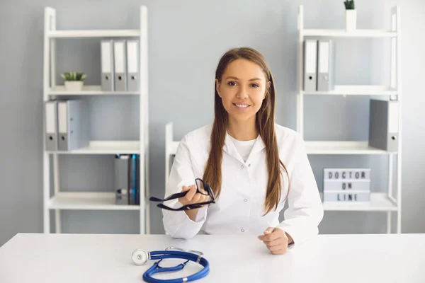 Médico sonriente mirando la cámara, consultando al paciente en línea en la clínica. GP de tener webcam videoconferencia con el cliente —  Fotos de Stock