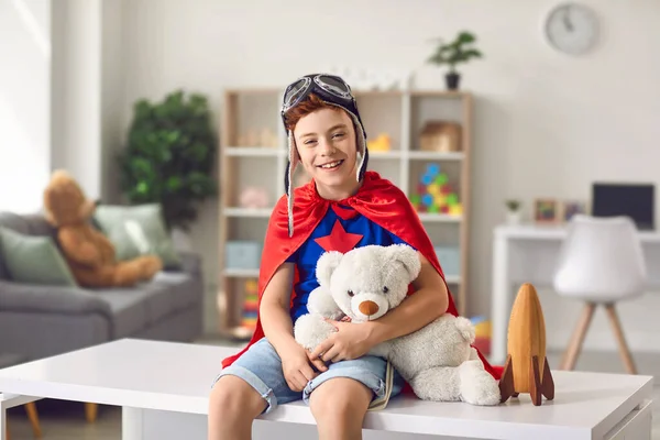 Menino em traje de super-herói sentado na mesa em casa com brinquedo e sorrindo — Fotografia de Stock