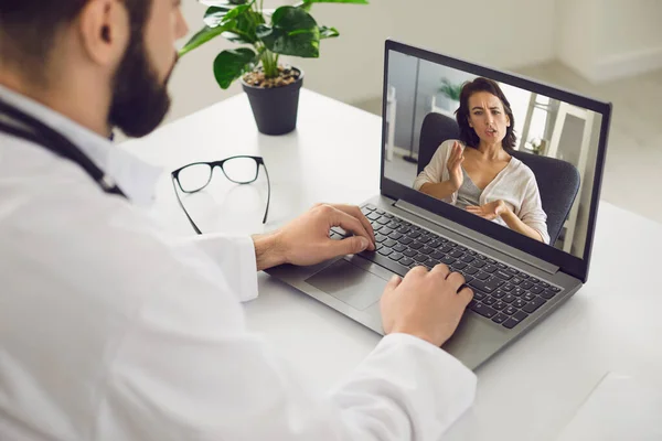 Online orvosi konzultáció. Egy férfi ül az irodájában használ egy laptopot, egy videohívást és hallgat egy beteget.. — Stock Fotó