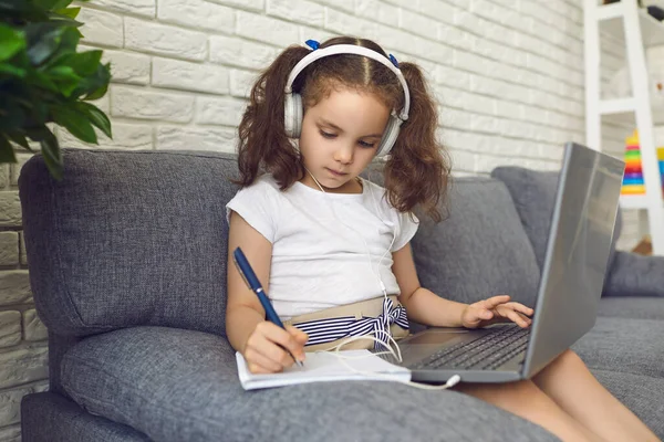 Niño de educación en línea. Niño adorable en auriculares viendo video lección en el ordenador portátil y tomando notas en casa —  Fotos de Stock