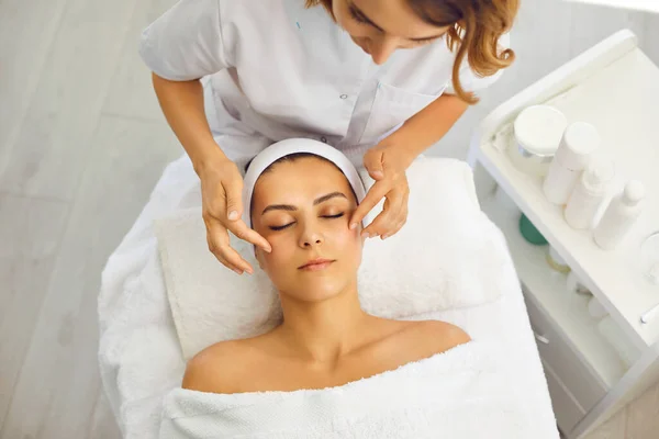 Cosmetólogo o dermatólogo poniendo crema durante el masaje facial para mujer en salón de belleza —  Fotos de Stock