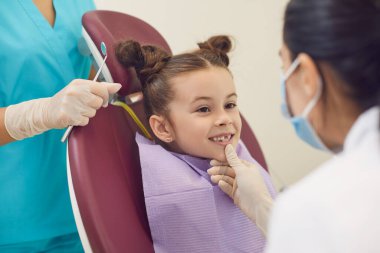 Kadın dişçi ve asistan, dişçi kliniğinde gülümseyen küçük kız dişlerini inceliyor.