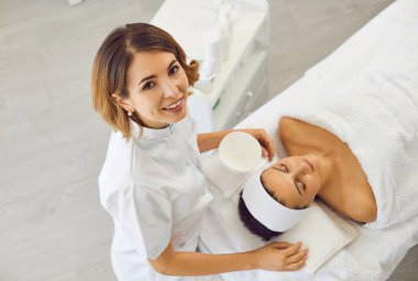 Kozmetik uzmanı ya da dermatolog güzellik salonundaki kadının yakınındaki kameraya bakıyor.