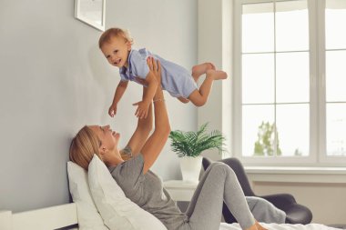 Mutlu anne kanepede uzanıyor ve kucağında gülümsüyor ve kaldırılmış ellerle kamera bebeğine bakıyor.