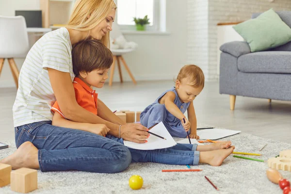 Młoda matka uczy dwójkę małych dzieci rysować kolorowymi ołówkami siedząc w pokoju. — Zdjęcie stockowe