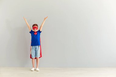 Süper kahraman kostümlü gülümseyen çocuk elleri gri arka planda duruyor.