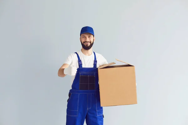 Trabalhador de entrega segurando uma caixa de papelão e mostrando polegares para cima em um fundo de parede cinza. — Fotografia de Stock