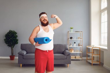 Spor kıyafetli komik şişman adam evde duran kaslarını gösteriyor.