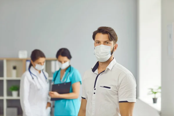 Man patient in medical protective mask standing over talking women doctors at background in clinic — Stock Photo, Image