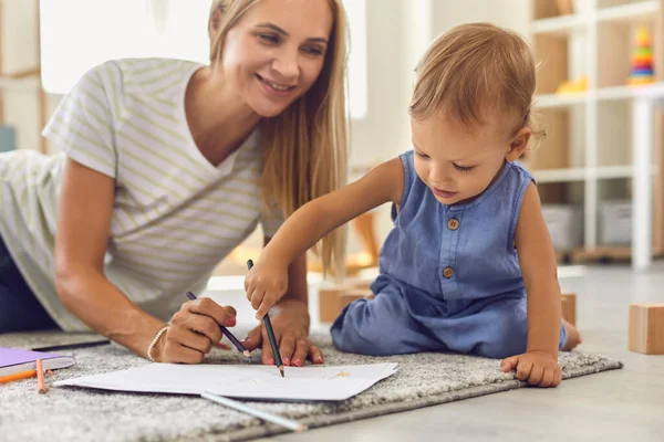 Bayi yang lucu menulis di atas kertas dengan pensil sambil duduk di lantai dengan pengasuhnya yang masih muda — Stok Foto