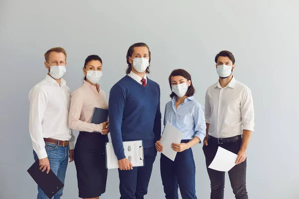Group of young positive business people in medical protective masks standing with documents in office