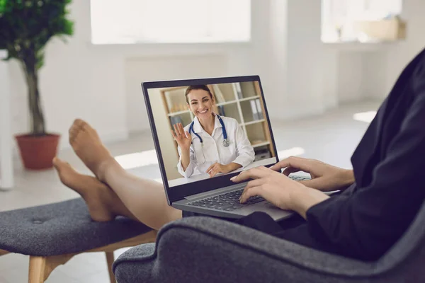 A young woman sits at home in a chair and calls the doctor online. Female doctor waves her hand to the patient. — Stock Photo, Image