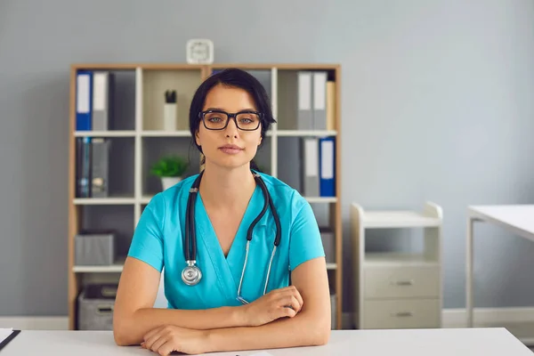 Terapeuta médica positiva sentada en el consultorio de la clínica médica y mirando la cámara durante la consulta en línea —  Fotos de Stock