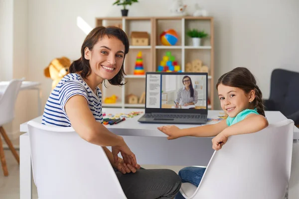 Улыбающиеся мама и дочь смотрят в камеру во время онлайн-урока с частным репетитором — стоковое фото