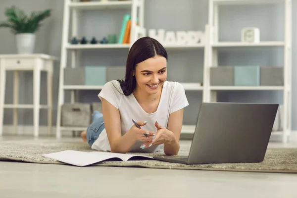 Lächelnde Frau, die auf dem Boden liegt, auf Laptop schaut und während des Online-Unterrichts etwas mit den Händen erklärt — Stockfoto
