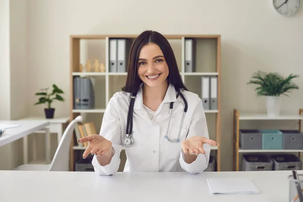 Mulher sorridente médico olhando para a câmera e mostrando algo com as mãos na clínica médica — Fotografia de Stock