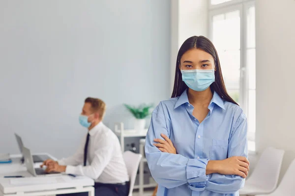 Wanita muda yang serius mengenakan masker wajah berdiri di tempat kerjanya sambil melihat kamera — Stok Foto