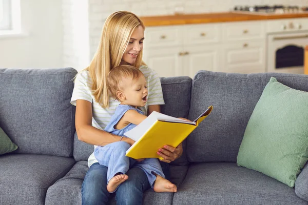 Młoda mamusia czyta bajki swojemu dziecku siedzącemu na kanapie w domu — Zdjęcie stockowe