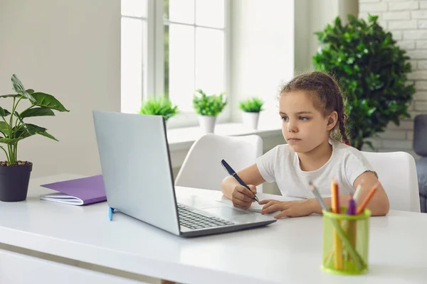 Інтернет-школи. Маленька дівчинка бере участь у відео уроці на ноутбуці вдома. Дитина робить нотатки під час вебінару вдома — стокове фото