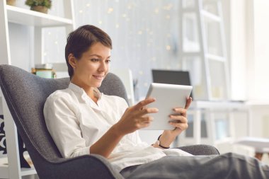 Tablette videolar izleyen mutlu bayan oturma odasındaki rahat koltukta oturuyor.