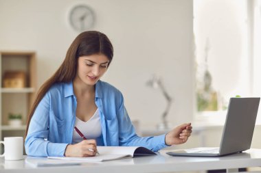 Evde dizüstü bilgisayarla oturup notlar alan genç bir kadın.