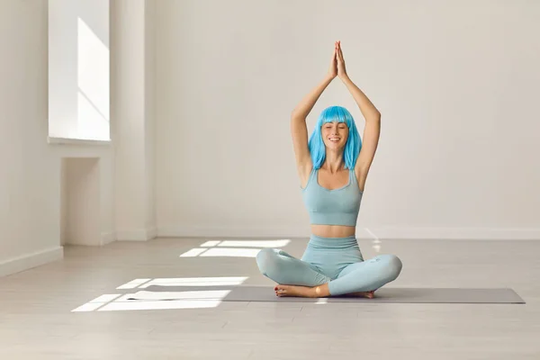 Młoda dziewczyna w stroju aktywnym siedzi z skrzyżowanymi nogami i ramionami podniesionymi na macie do jogi w nowoczesnej siłowni — Zdjęcie stockowe