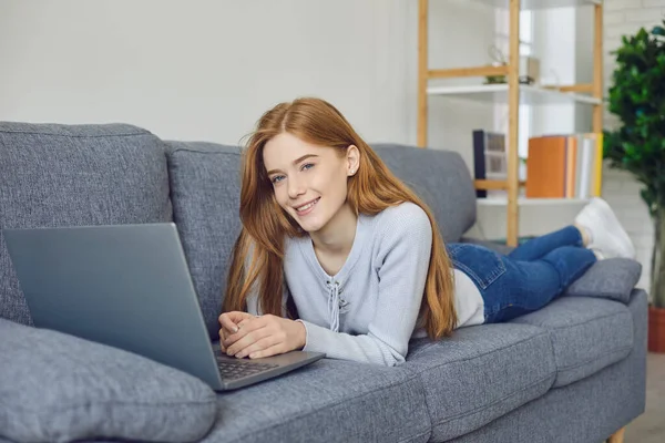 Lächelndes Mädchen, das mit Laptop auf dem Sofa liegt und zu Hause in die Kamera schaut — Stockfoto