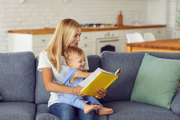 Ibu yang bahagia menggendong anaknya yang masih bayi berlutut dan membaca buku bersama di sofa — Stok Foto