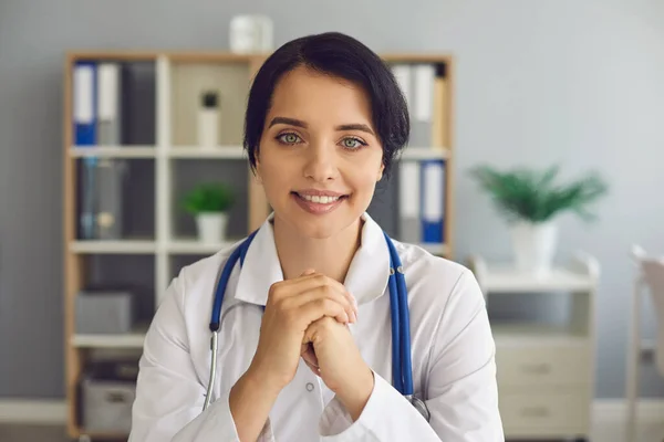 Amichevole giovane medico guardando la fotocamera durante la consultazione online nel suo moderno ufficio ospedaliero — Foto Stock