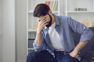 Üzgün adam evde oturuyor, kötü hissediyor ve elleriyle yüzüne dokunuyor.