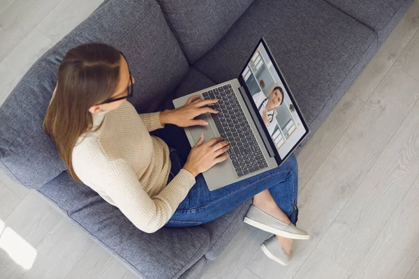 Family doctor online. Young girl at home on sofa with laptop makes video call to doctor. Top view.