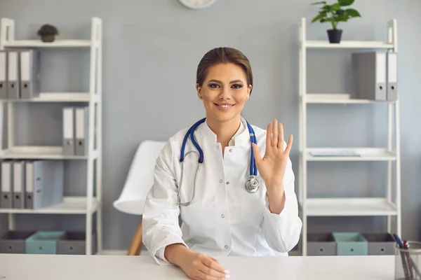 긍정적 인 의사가 병원 사무실 테이블에 앉아서 카메라를 보고 있는 환자에게 손을 흔들고 있습니다.. — 스톡 사진