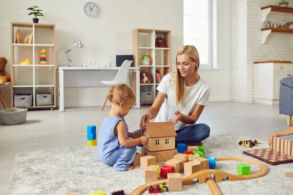 Joven mamá jugando con lindo hijo pequeño y enseñándole a construir una casa de juguete en un acogedor apartamento moderno —  Fotos de Stock