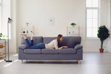 Genç, gülümseyen bir kadın evdeki kanepede yatıyor ve internette dizüstü bilgisayarla çalışıyor.